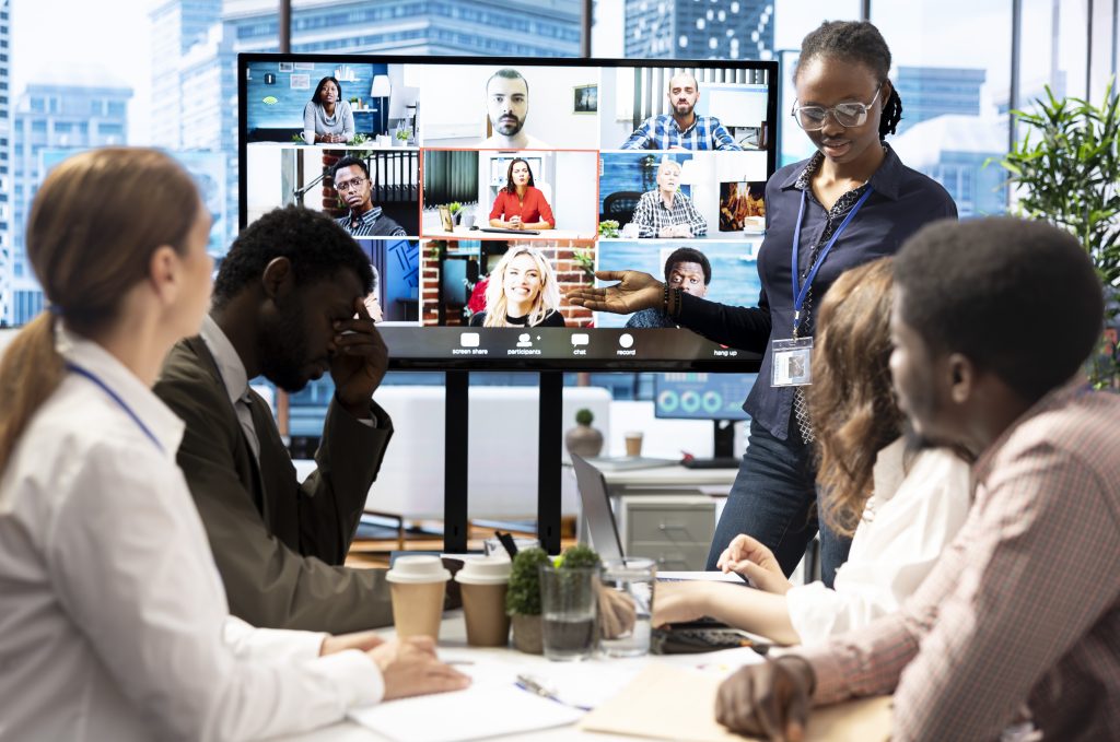 Hybrid team meeting with five people around a conference room table and nine people on a large screen
